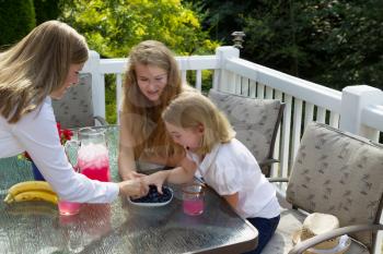 Side view of happy mother and daughters reaching for fresh blue berries while outdoors on patio with woods in background 
