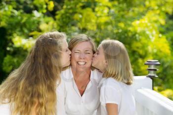 Front view of mother, eyes closed, while being kissed by her two daughters outdoors on patio with blurred out woods in background 