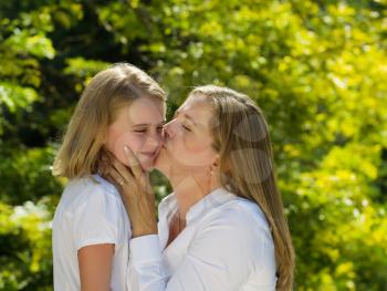 Side view of mother kissing her youngest daughter while holding her face in her hands during outing outdoors on patio with blurred out woods in background 
