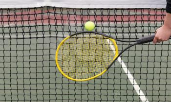 Horizontal photo of a tennis racket in front of ball with net and court in background