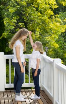 Vertical side view of younger sister measuring height difference with older sister while outdoors on patio with blurred out trees in background 