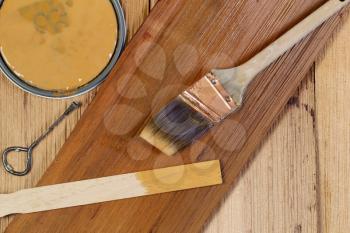 Closeup top view of painting tools consisting of hand brush, stir stick, can opener, and paint lid on cedar wooden shingles with top board stained and unstained boards underneath 