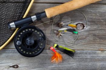 Close up top view of a fishing fly reel, with line, landing net and assorted flies, and partial cork handled pole on rustic wooden boards 

