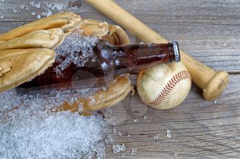 Close up of a brown bottled beer with crushed ice inside of baseball glove on rustic wood with ball and bat 

