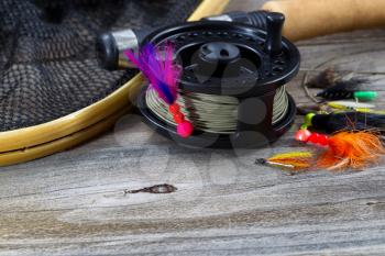 Close up of fly reel, focus on front of reel, with fly jig hanging from spool. Partial cork handled pole, net and flies blurred out on rustic wooden boards 