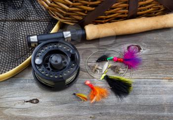 Close up top view of  fishing fly reel, landing net, creel and assorted flies, with partial cork handled pole on rustic wooden boards 