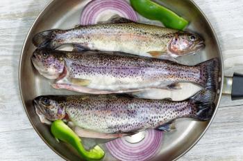 Top view of fresh wild trout in frying pan with onion and green peppers on rustic white wood. 
