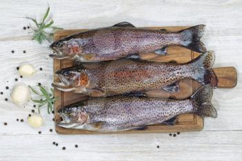 Top view of fresh trout being prepared, skin coated with oil, for cooking on server board. Herbs and spices on white aged wooden boards. 