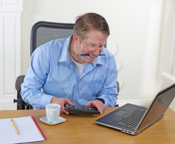 Mature man biting pencil while looking at laptop screen with calculator, pencils, coffee cup and notepad on desk. Background is white walls.  