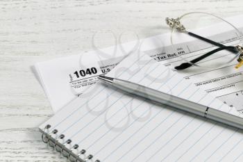 Close up of a silver business pen on top of notepad with tax form and reading glasses in background on white desk. Focus on tip of pen. 
