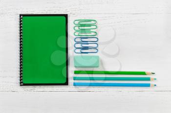Green spiral notepad with pencils, eraser, and paper clips on desktop. Pattern in colors of green and blue with a white wooden background.  Educational or business concept. 
