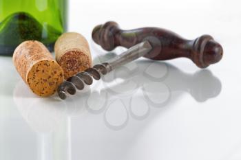 Close up front view of antique corkscrew, focus on front part of corkscrew and cork on far left side, on glass table with green empty wine bottle in background along with reflections 