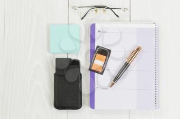 Angled top view image of a business desktop consisting of the following items: calendar, pen, reading glasses, paper stickers and cell phone inside of case on white wood. 