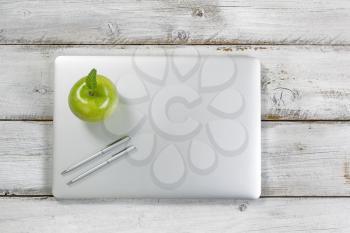 Green apple and silver pens on top of laptop with rustic desktop in background.  