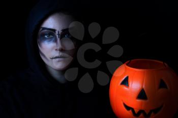 Teen girl in scary face paint holding large pumpkin container on black background. Trick or treat concept for Halloween with selective lighting.  