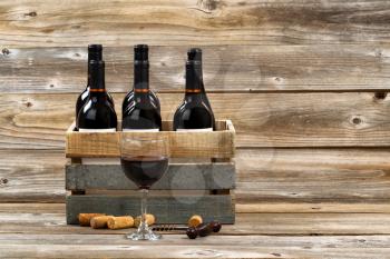 Glass of red wine with full bottles in wood crate, old corkscrew and used corks on rustic wooden boards. 
