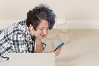 Smiling teen girl looking at cell phone with computer in forefront while lying down listening to music at home. 