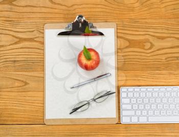 Top view of clipboard, computer keyboard, reading glasses, apple, paper and pen on desktop. 