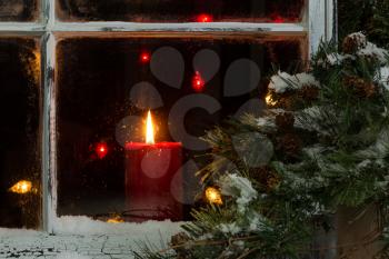 Close up of a red candle, selective focus on flame and top part of candle, glowing in window with pine tree and snow outside. Christmas concept.