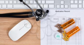 Patient medication record form with stethoscope, medication container, capsules, computer keyboard and mouse on wooden desktop. Mouse and keyboard are generic brand items for display purposes only. 