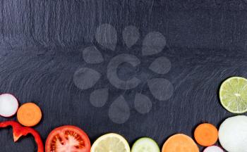 Overhead view of various sliced vegetables and fruits, on bottom of frame, on natural slate stone. 