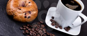 Angled view of roasted coffee beans on natural black slate stone with coffee drink and bagel in background. Selective focus on coffee beans in forefront. 