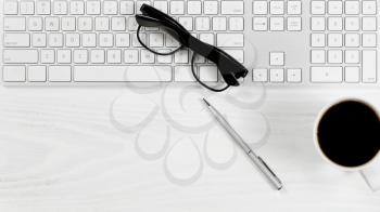 Overhead view of a white desktop with keyboard, coffee, pen and reading glasses. 
