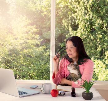 Mature woman, wearing pink bathrobe, holding her family cat while working from home in front of large window with bright daylight and trees in background. Haze light effect applied to image. 