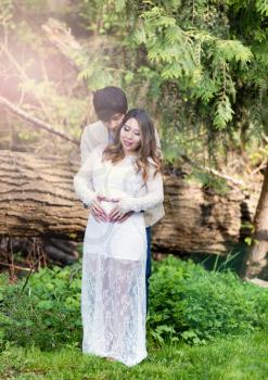 Expecting mom and dad holding each other with heart shaped hands over her abdomen with woods in background. Haze lighting effect on left side of image.