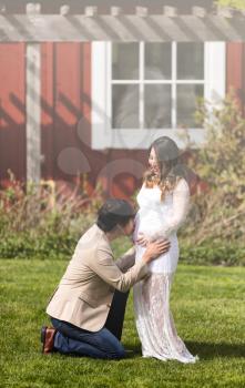 Husband kissing abdomen of expecting mom while kneeling in front of her on grass. Haze light effect applied to image. 