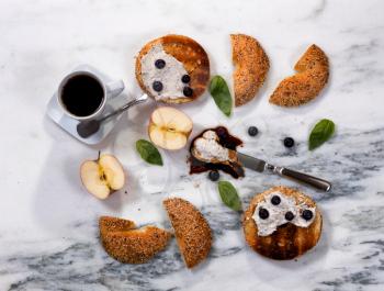 Overhead view of freshly toasted bagels with cream cheese, sauce, blueberries, basil, sliced apple and coffee on white marble stone. Horizontal format. 