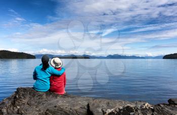 Daughter and mother, facing away from camera, enjoying the outdoors on a beautiful day with lake and mountains in background.  Selective focus on backs of people. 