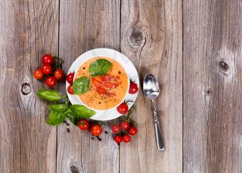 Overhead view of fresh creamy tomato soup, in white bowl, with cherry tomatoes and basil on rustic wooden boards. 