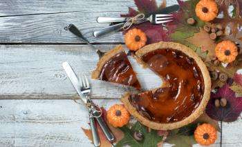 A slice of pumpkin pie with autumn foliage and small pumpkins on rustic white wood.  Thanksgiving holiday concept in flat lay format. 
