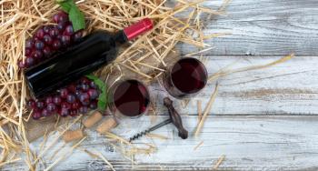 Overhead view of a red wine bottle, drinking glasses, grapes plus corkscrew with straw and burlap on white rustic boards 