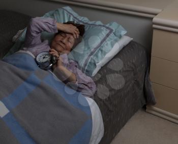 senior woman with painful headache holding her alarm clock during nighttime