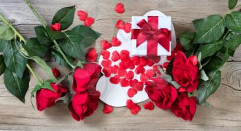 red roses surrounding white gift box and many hearts on weathered wooden background in flat lay view 