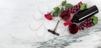 Overhead view of Red wine bottle with grapes, drinking glasses, corkscrew and rose on natural marble stone