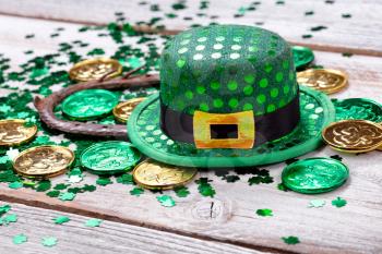 Bright green hat with gold coins, shamrock and rusty horseshoe for Saint Patrick Day on white rustic wooden background in close up format  