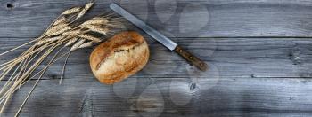 Top view of homemade freshly baked loaf of bread with cutting knife and dried wheat stalks on weathered wooden planks 