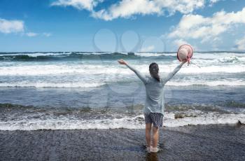 Woman facing ocean and blue sky with arms up while holding her hat in expression of pure joy                               