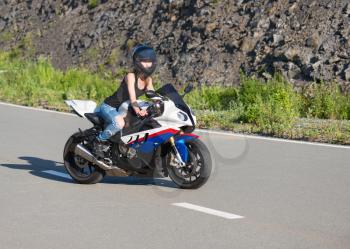 Young beautiful blonde girl in trendy blue jeans and a black T-shirt on the nature behind the wheel of modern motorcycle. Portrait in soft sunny colors.