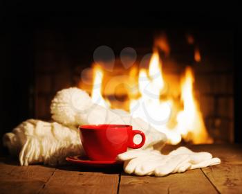 Red cup of coffee or tea, woolen scarf, gloves and cap on wooden table near  fireplace. Winter and Christmas holiday concept. 