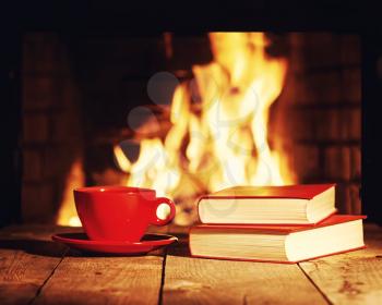 Red cup of tea or coffee and old books near fireplace on wooden table. Winter and Christmas holiday concept. Photo with retro filter effect.
