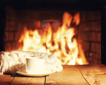 White cup of tea or coffee and woolen scarf near fireplace on wooden table. Winter and Christmas holiday concept. Photo with retro filter effect.