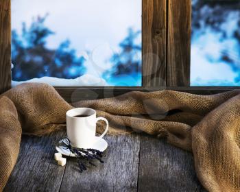 White cup of coffee or tea, lavender flowers and natural gunny cloth located on stylized wooden window sill. Winter concept of comfort and relaxation.