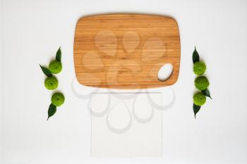 Wooden cutting board and paper with decoration of chrysanthemum flowers and ficus leaves on white background. Overhead view. Flat lay.