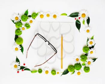 Paper, pencil and glasses with wreath frame from chamomile and chrysanthemum flowers, ficus leaves and ripe rowan on white background. Overhead view. Flat lay.
