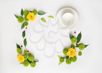 Cup for coffee or tea with wreath frame from roses, chamomile and chrysanthemum flowers, ficus leaves and ripe rowan on white background. Overhead view. Flat lay.