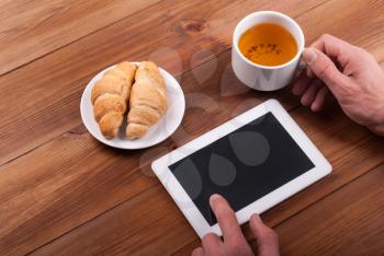 Hands of a man with a digital tablet and a cup of tea on a wooden table.